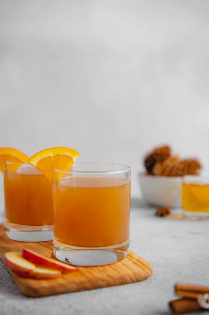 Straight on photo of two glasses of apple cider with pinecones and a shot glass in the background.