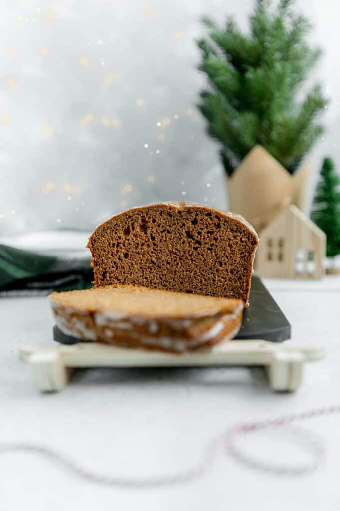 Straight on photo of the inside of a gingerbread loaf with a slices cut out in the foreground and a Christmas tree and plates in the background.