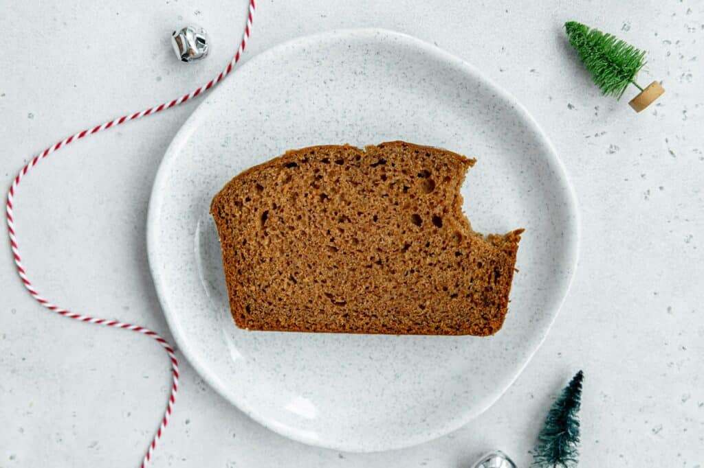 Overhead photo of a slice of a gingerbread loaf on a plate and a couple mini bottle brush trees and jingle bells.