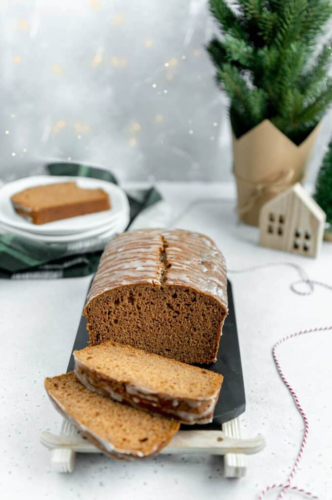 Photo of a gingerbread loaf with a couple slices cut and a Christmas tree and plates in the background.