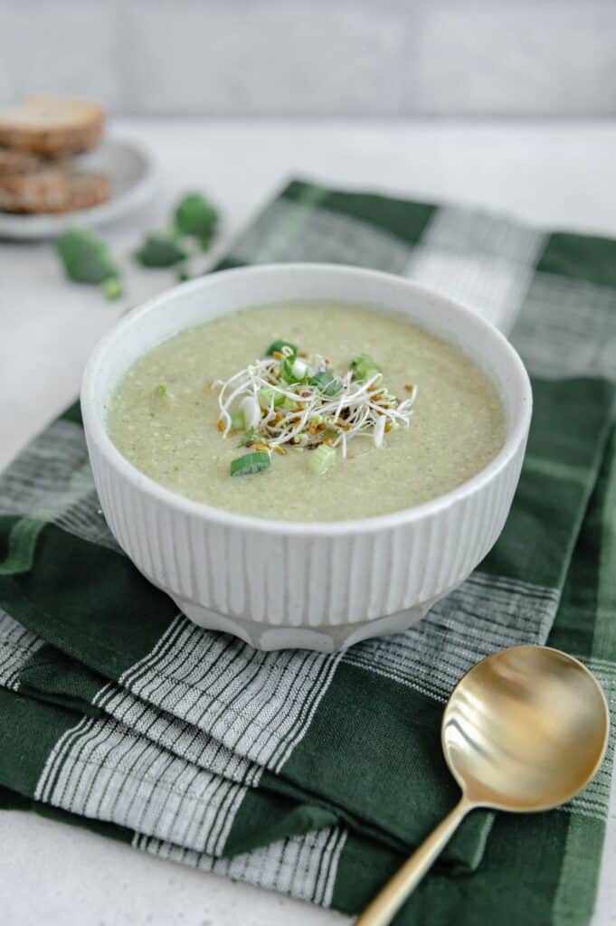 45-degree photo of a bowl of broccoli soup with a gold spoon in the foreground and stacked bread sliced in the background.