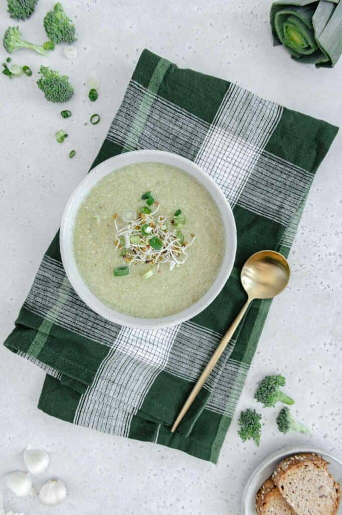 Overhead photo of broccoli soup bowl on top of a napkin with broccoli, leeks, garlic and bread around the edges of the photo.