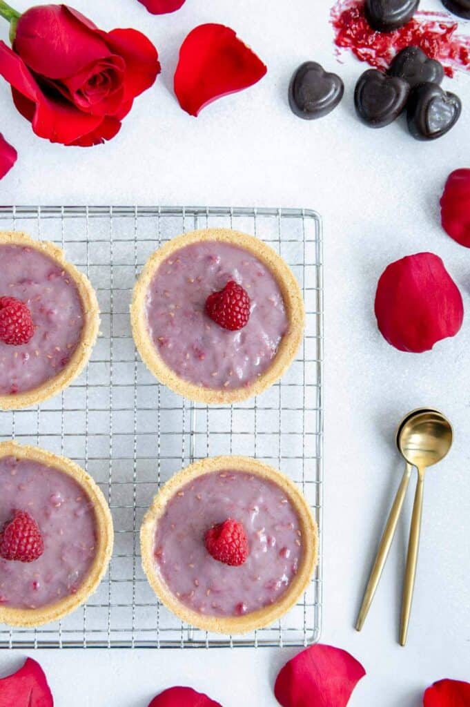 For raspberry tarts on a cooling rack with chocolate and rose petals scattered around.