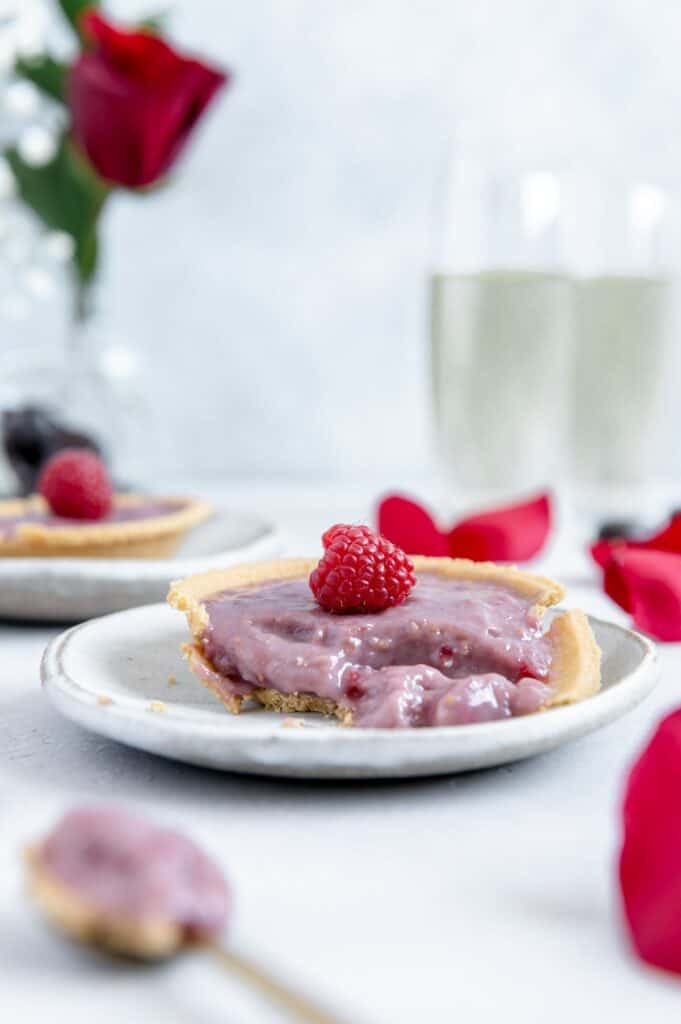 Half eaten raspberry tart on a plate with a rose and wine glasses in the background.