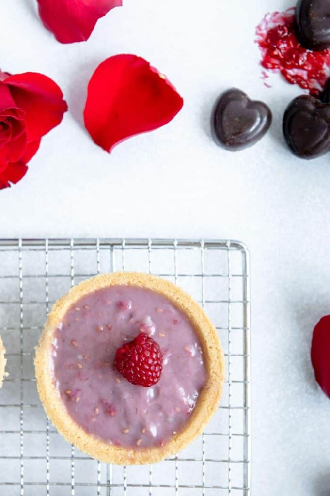 Raspberry tart on a cooling rack with chocolate and rose petals scattered around.