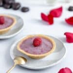 Raspberry tart on a plate with a spoon, and wine glasses, rose petals and chocolate in the background.