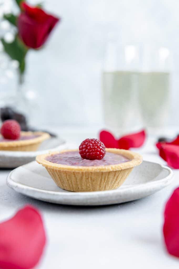 Raspberry tart on a plate with a rose and wine glasses in the background.