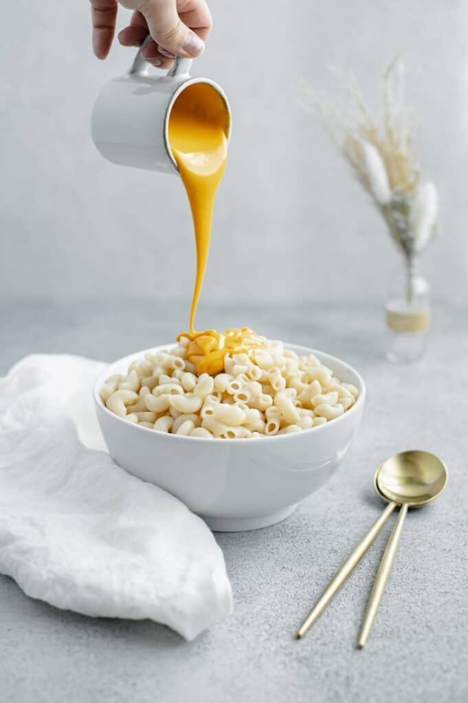 A hand pouring cheese sauce overtop a bowl of macaroni.