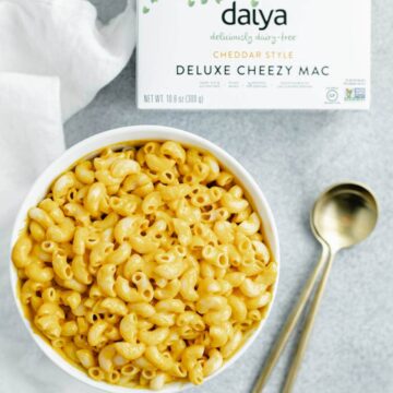 A bowl of mac and cheese with two spoons next to it and a box of Daiya deluxe cheesy mac.
