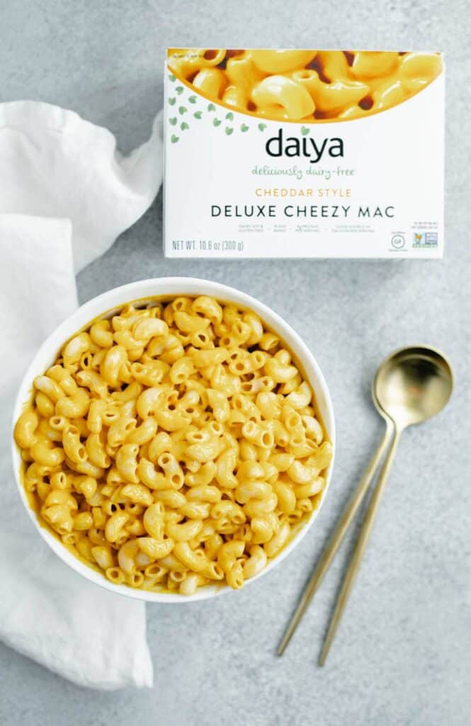 A bowl of mac and cheese with two spoons next to it and a box of Daiya deluxe cheesy mac.