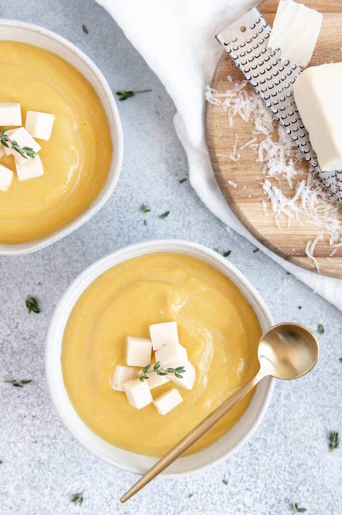 Overhead of two pumpkin soup bowls with cheese on top and cheese on cutting board with a cheese grater.