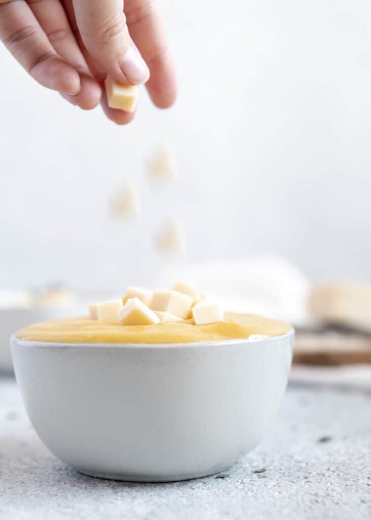 Hands dropping cubed cheese into a bowl of pumpkin soup.