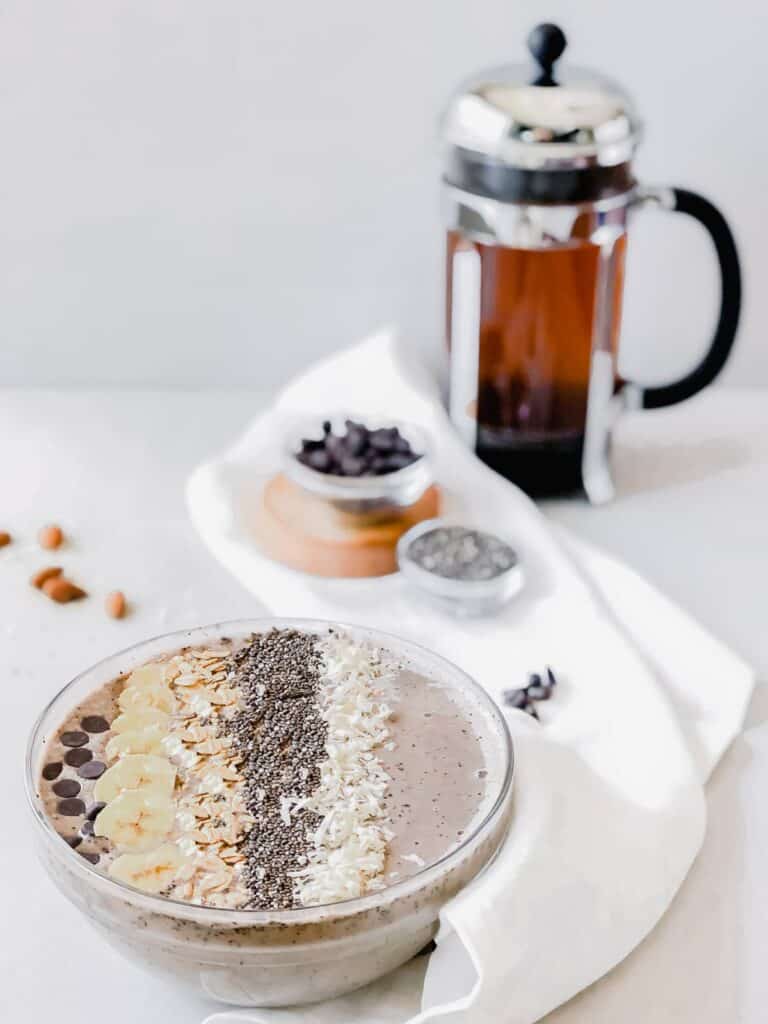 Coffee banana smoothie bowl with a coffee press and ingredients blurred in the background.