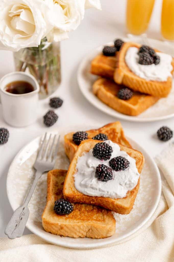 Three vegan French toasts on a plate with a fork next to them and flowers and mimosas in the background.