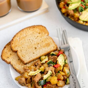 Vegan breakfast hash on a plate with toast and a fork and knife with two cups of coffee in the background.
