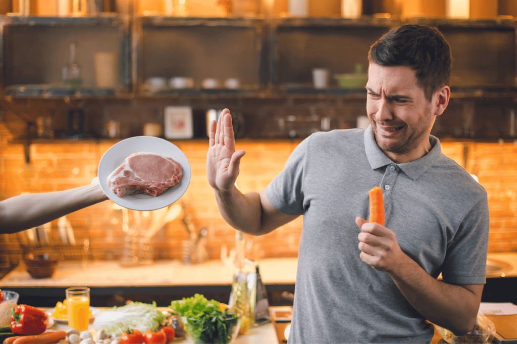 A man holding a carrot and saying no to a plate of meat.
