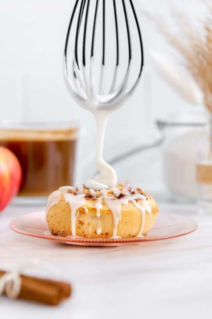 A whisk drizzling icing over top of a cinnamon roll.