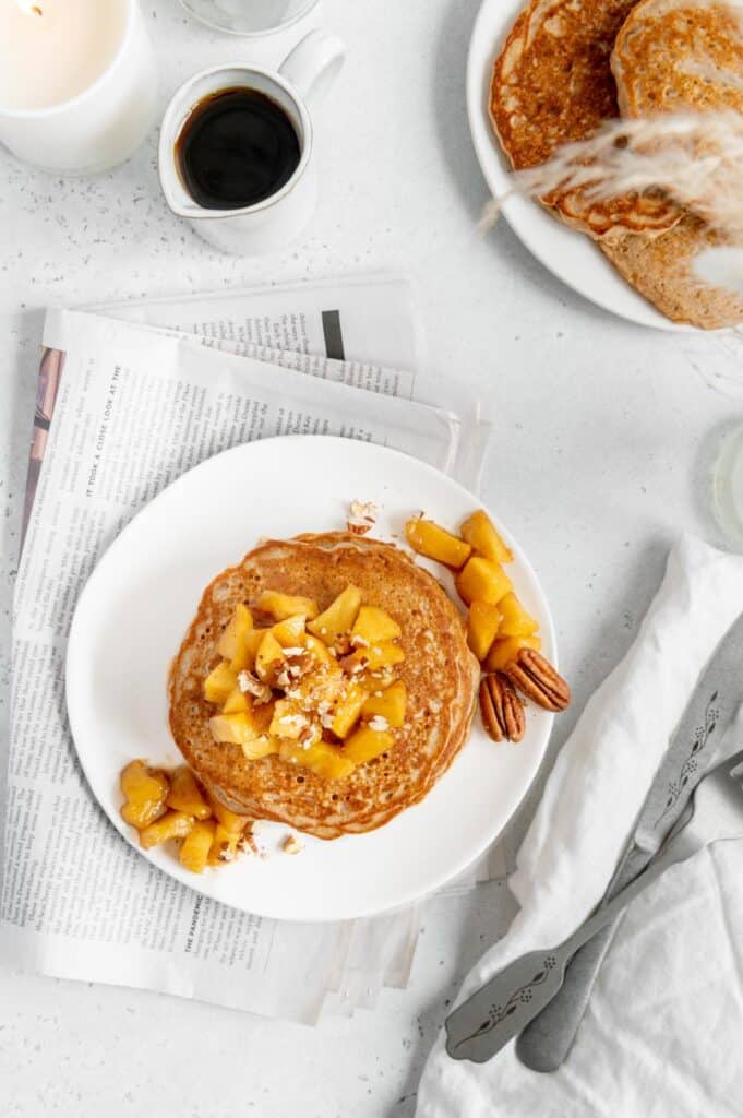A breakfast scene of vegan apple pancakes, newspaper, maple syrup, and a candle.