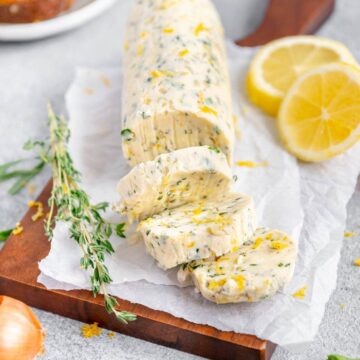A stick of vegan compound butter on a cutting board with lemons, herbs, and shallots scattered around.