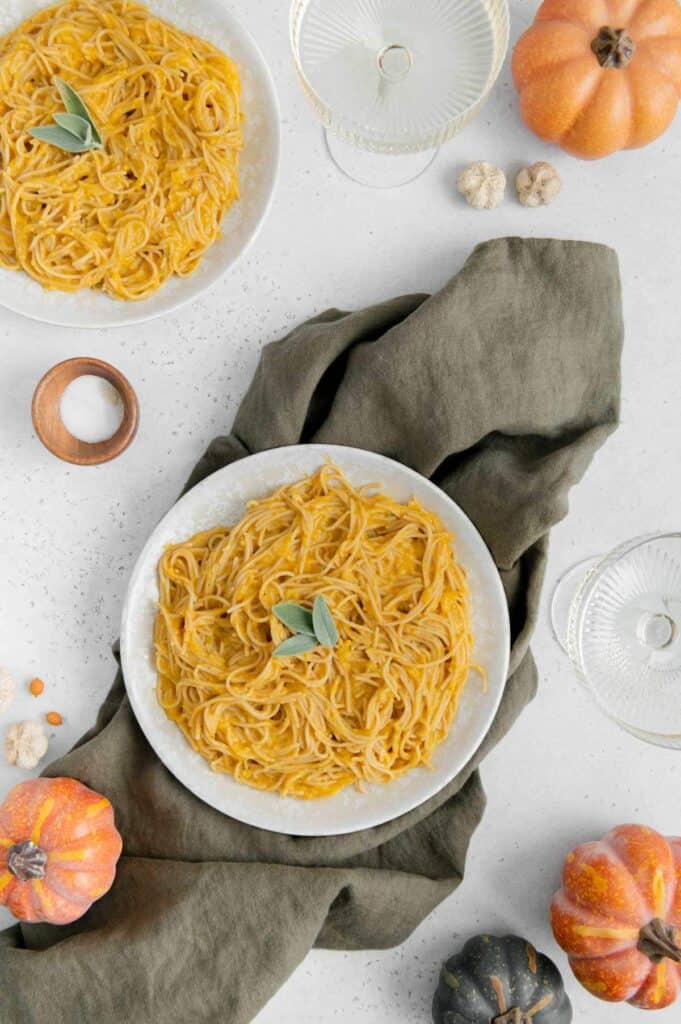 Two plates of vegan pumpkin pasta with wine glasses and pumpkins scattered around.