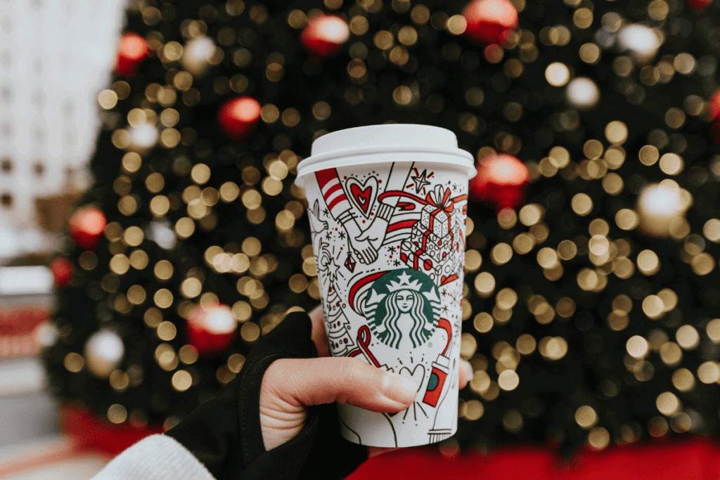 A holiday Starbucks cup in front of a Christmas tree.