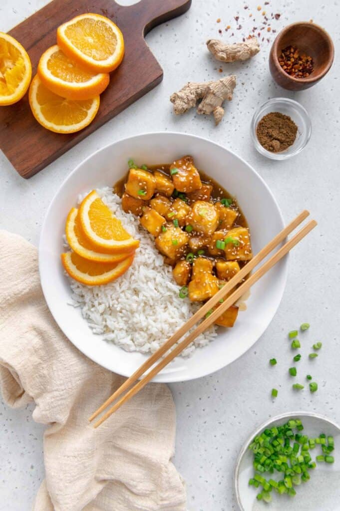 Chopsticks over a bowl of rice and vegan orange tofu.