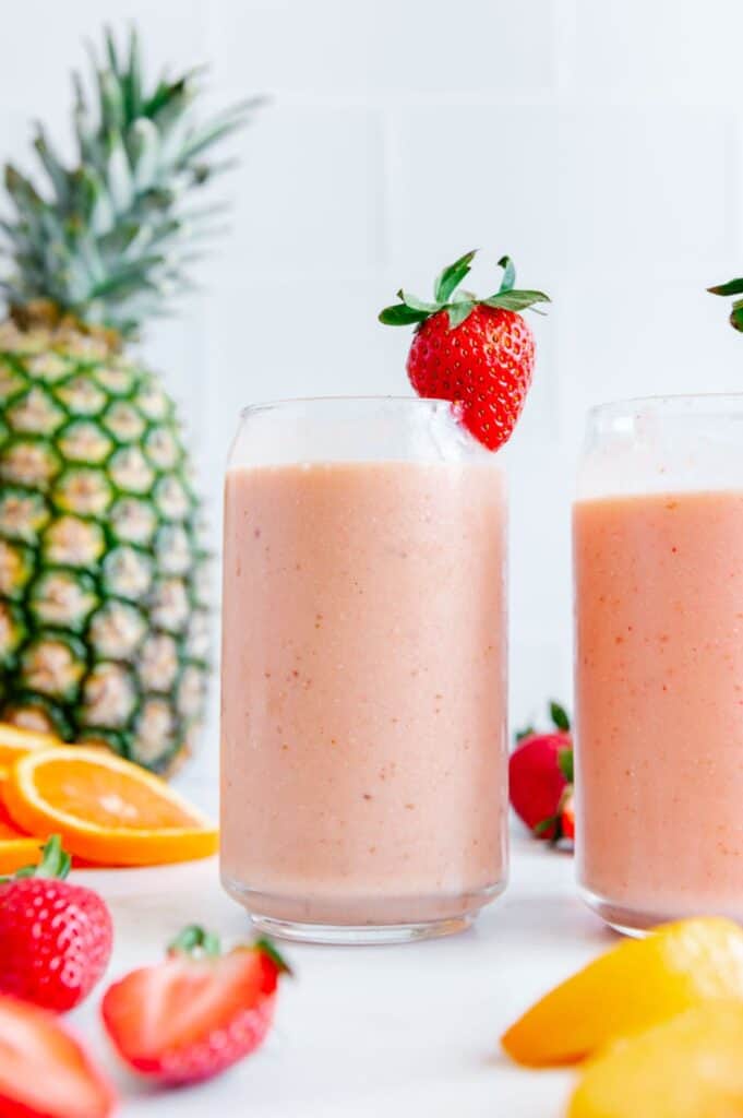 Two tropical smoothies surrounded by a lot of fresh fruit.