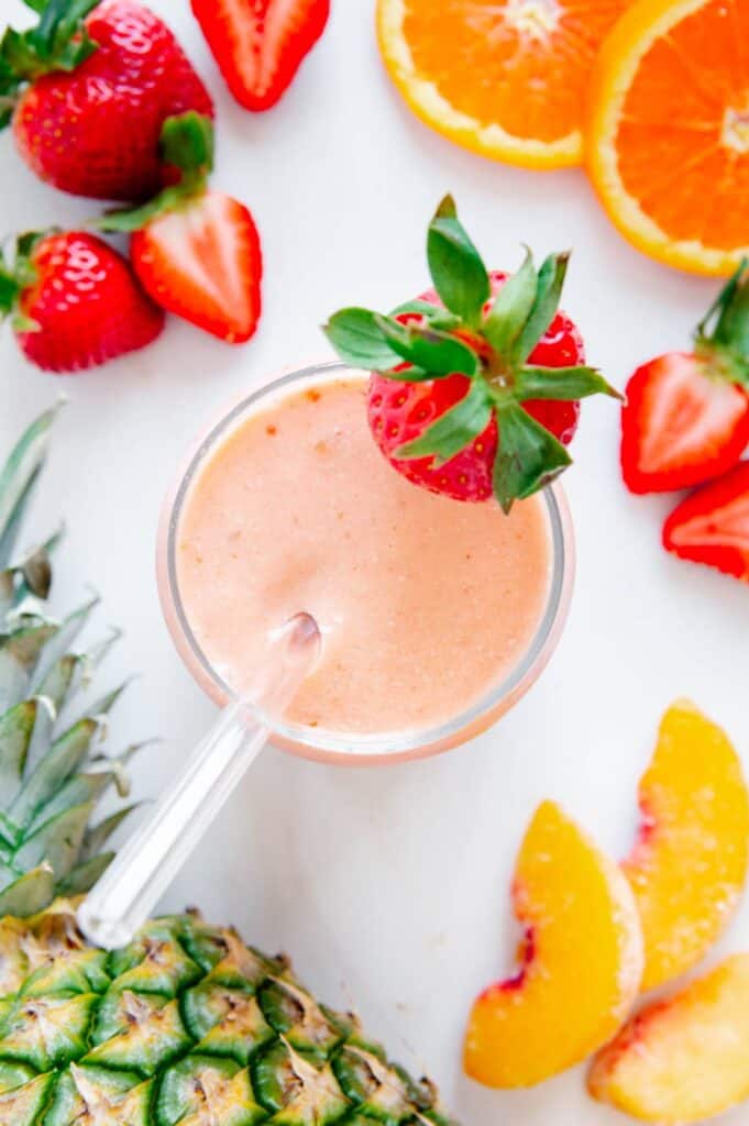 Overhead of a tropical smoothie surrounded by fresh strawberries, pineapple, oranges, and peaches.