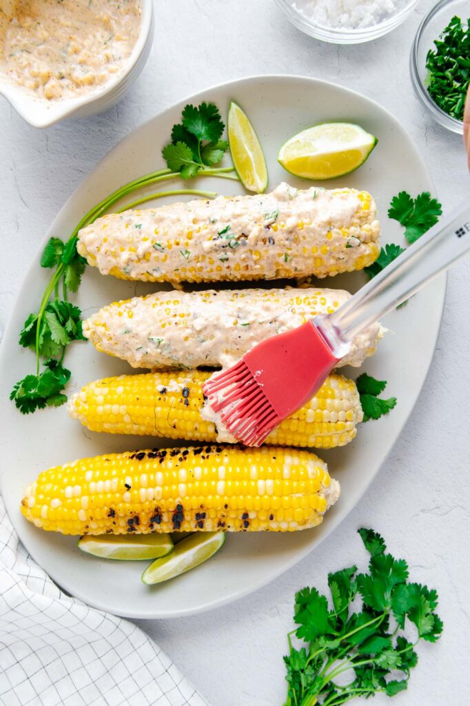 A silicone brush spreading Mexican crema over grilled corn on the cob.