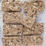 Eight healthy vegan granola bars surrounded by oats, nut-butter, and mini chocolate chips.