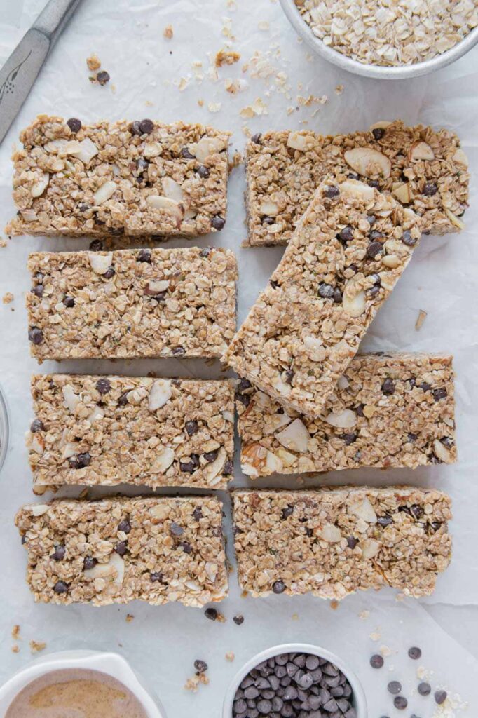 Eight healthy vegan granola bars surrounded by oats, nut-butter, and mini chocolate chips.