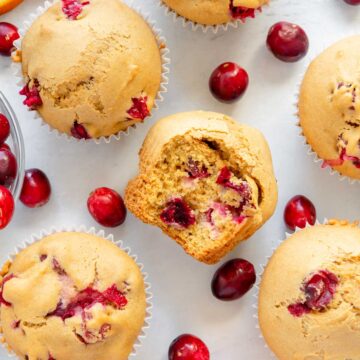 Scattered muffins and cranberries with a muffin in the center with a bite taken out of it.