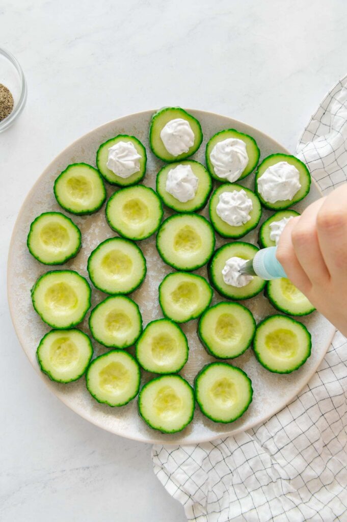 Vegan cream cheese and sour cream with herbs being piped onto cucumber slices.