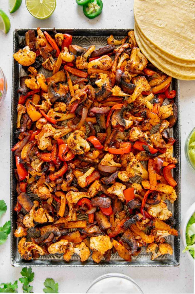 Sheet pan fajita veggies in spice mixture on a baking sheet surrounded by topping options.