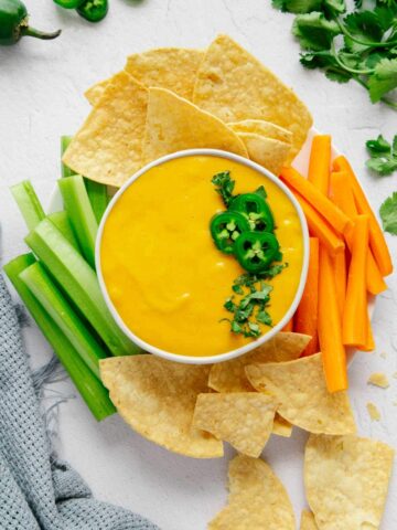 A bowl of vegan nacho cheese without cashews on a serving plate with chips, carrots, and celery.