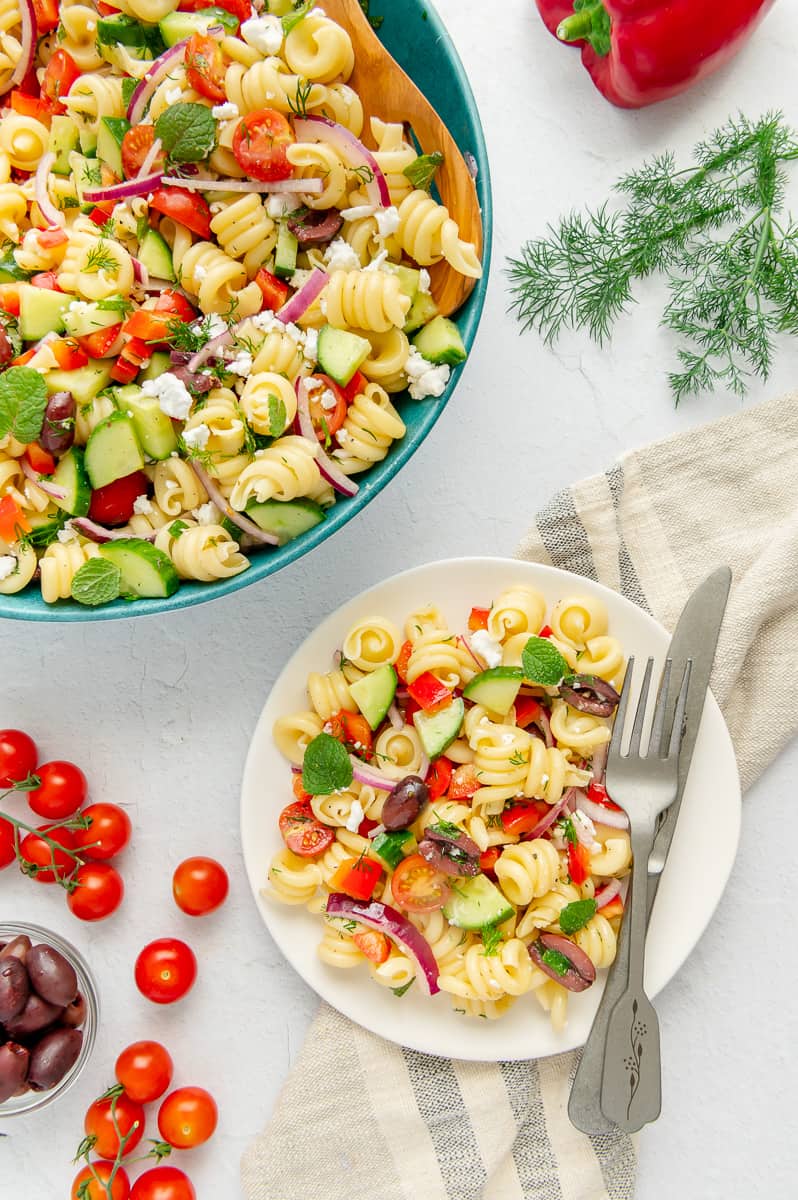 A white plate with a serving of Mediterranean pasta salad next to a fork and knife.