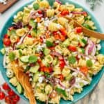 Mediterranean pasta salad with fresh veggies and herbs in a blue serving bowl with wooden serving spoons.