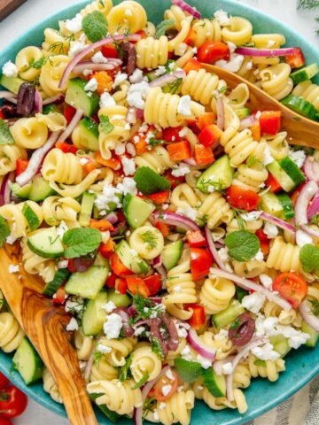 Mediterranean pasta salad with fresh veggies and herbs in a blue serving bowl with wooden serving spoons.