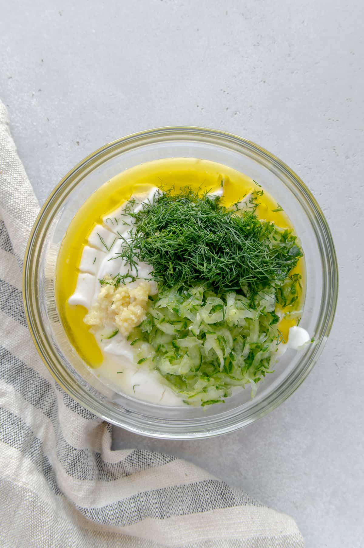 Vegan Greek yogurt, fresh dill, olive oil, lemon juice, cucumber and garlic in a mixing bowl.